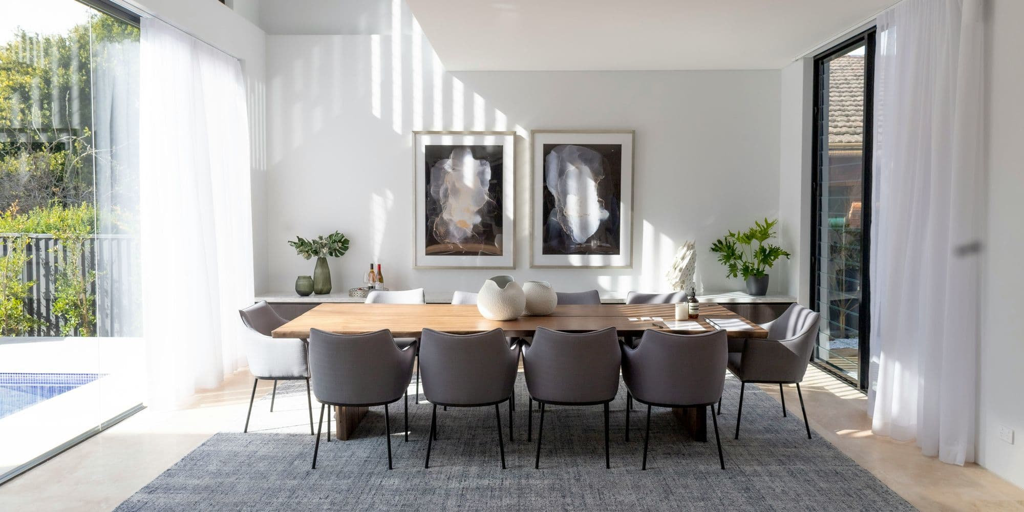 Sun-filled dining room with floor-to-ceiling windows, walnut table, and statement artwork on white walls